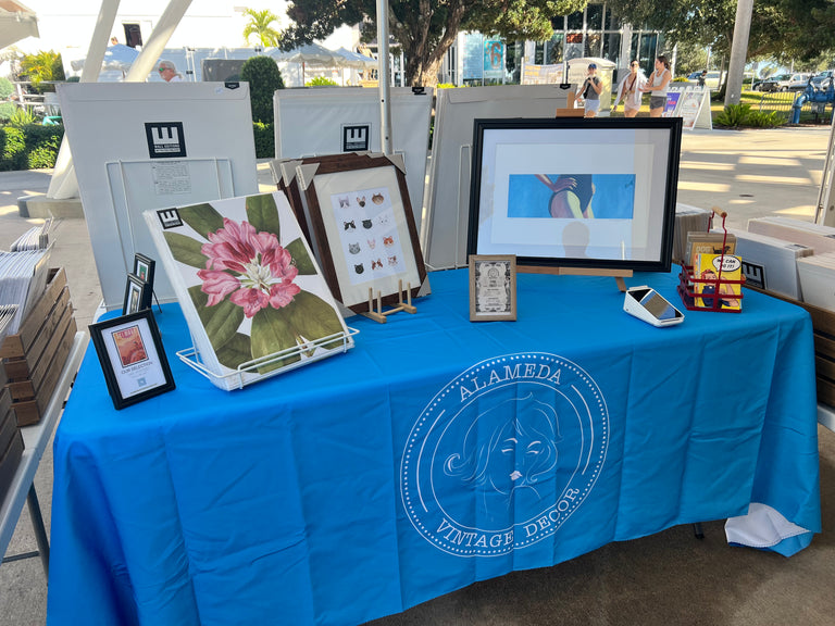 An outdoor booth display for 'Alameda Vintage Decor' featuring framed vintage-style prints, a pink flower artwork, and a small display box on a bright blue branded tablecloth.