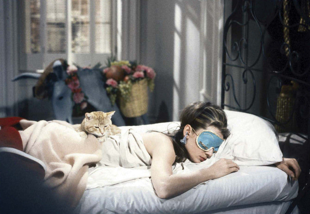 Woman lying on a bed with a cat, wearing a sleep mask, in a softly lit room.